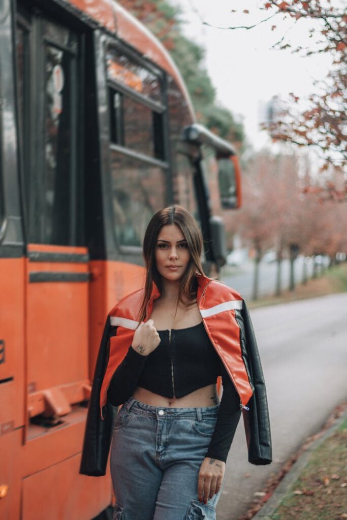 A stylish woman stands confidently by an orange bus in a fall setting.